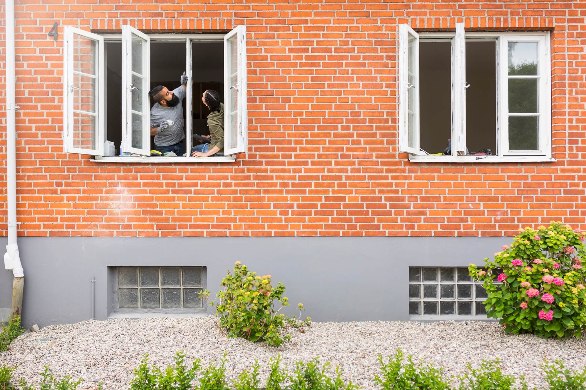 Två personer som arbetar med fönstermontering från insidan av ett öppet fönster på ett hus med tegelfasad.